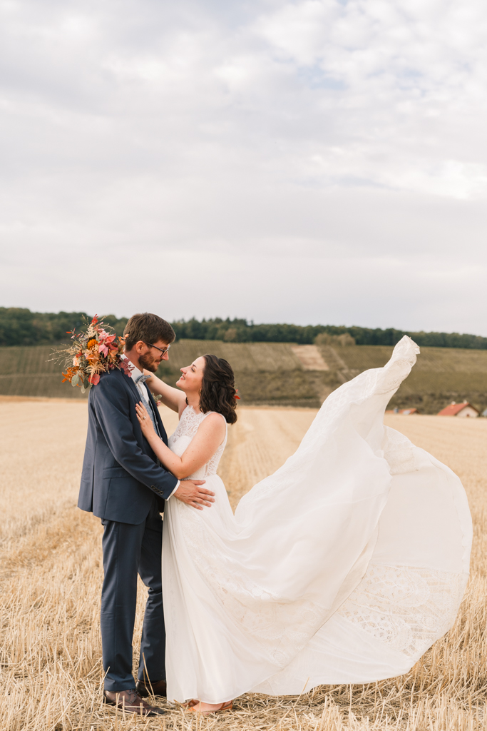 Hochzeitspaar steht auf einem Feld und die Schleppe fliegt im Wind.