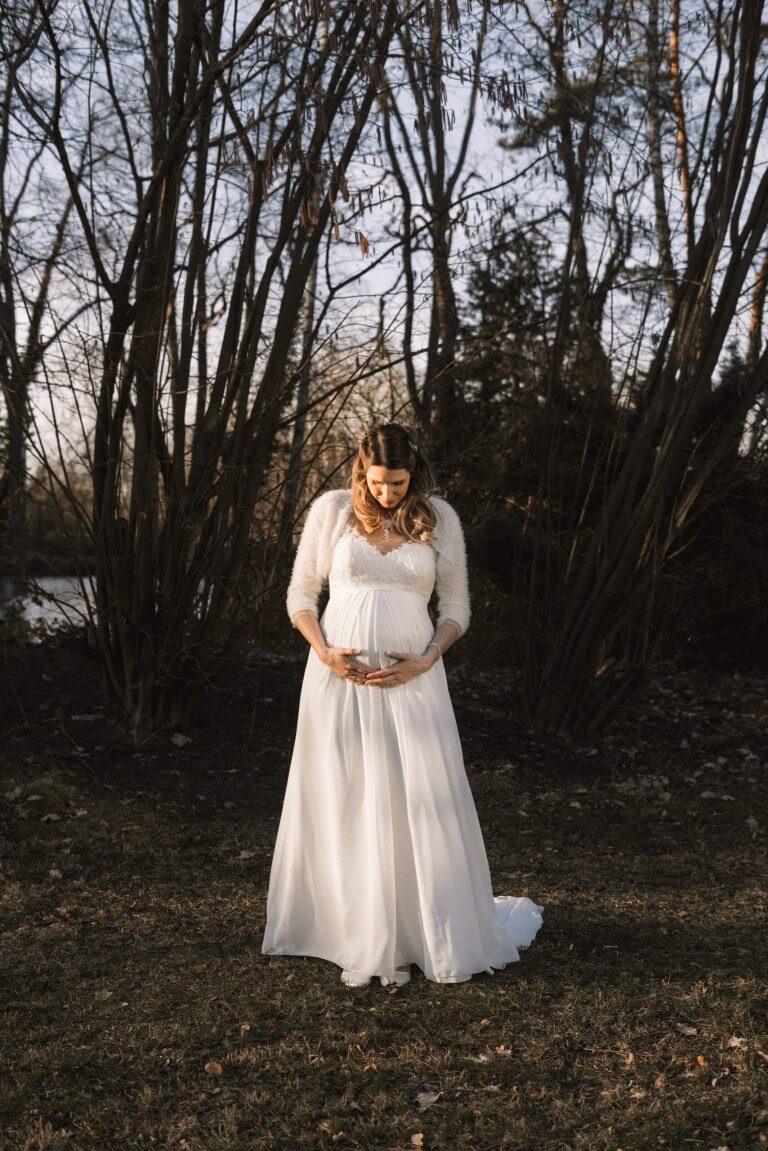 Braut steht bei untergehenden der Sonne draußen und hält unter ihrem Brautkleid den Babybauch in den Händen